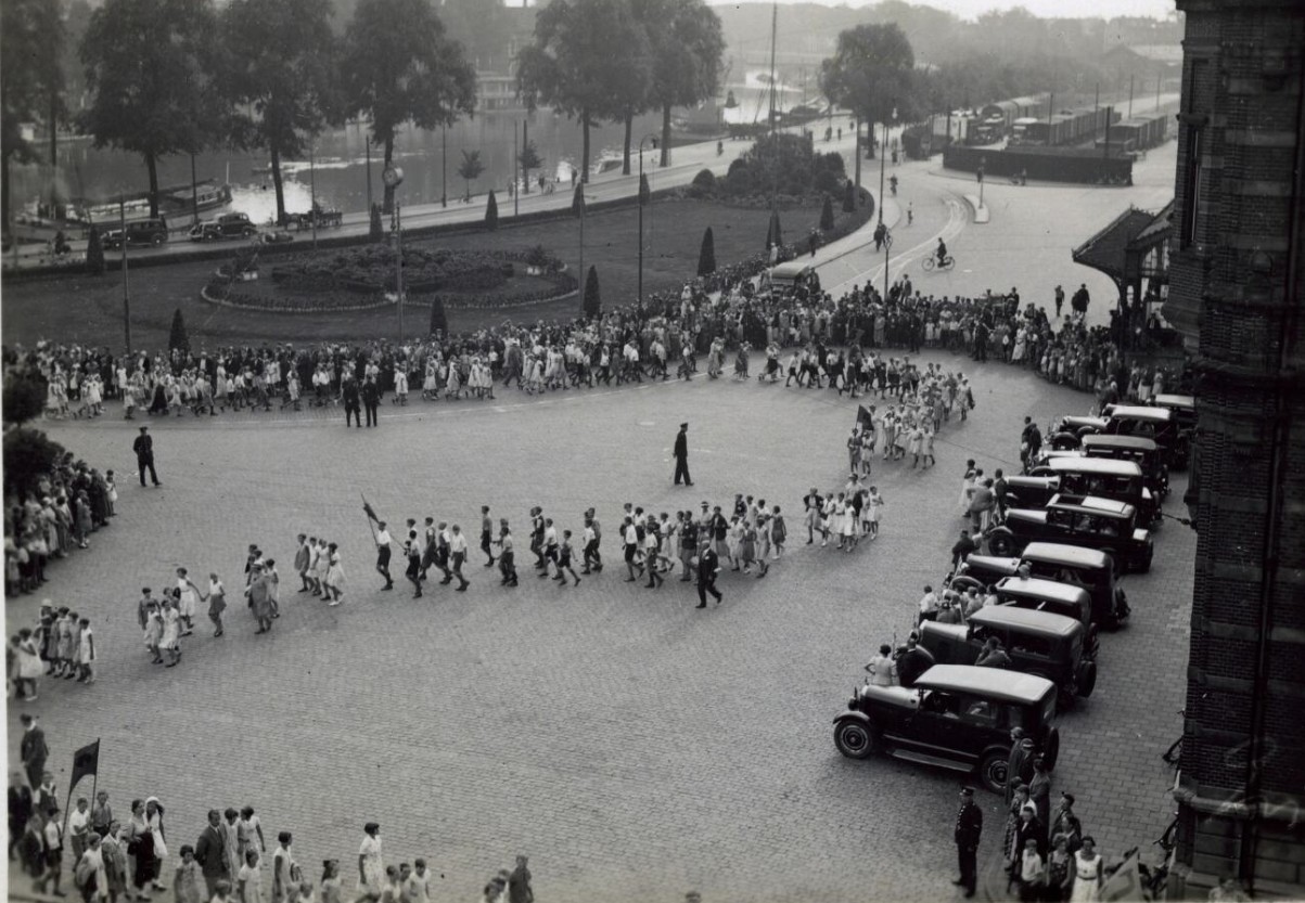 De vakantiekinderfeest stoet nadert het Hoofdstation in Groningen in 1934. In de auto's rechts werden de 'zwakke kinderen' vervoerd die een aangepast programma kregen.